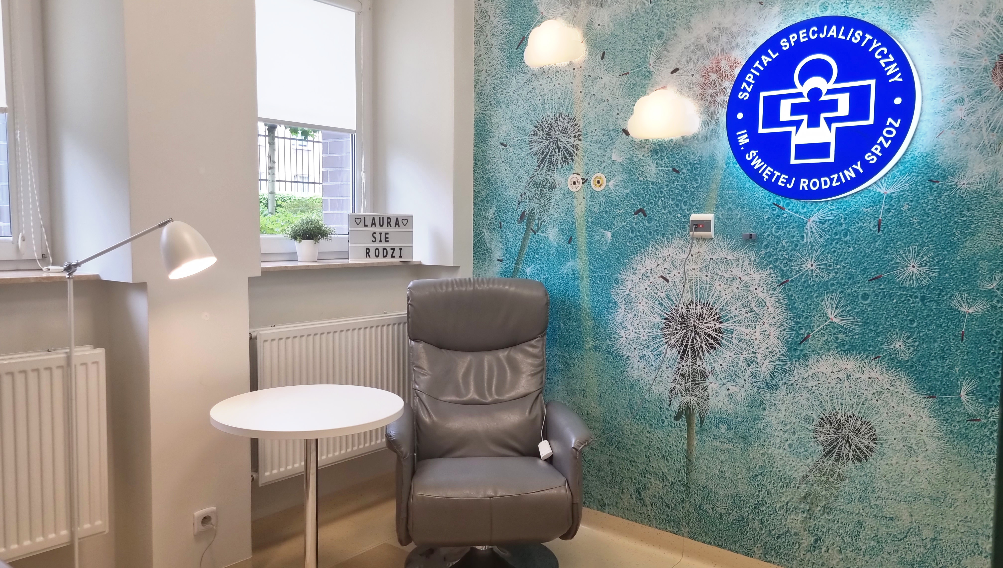 Przytulny kącik w szpitalu, z wygodnym fotelem, stolikiem, lampą i ścianą ozdobioną dmuchawcami. Idealne miejsce do relaksu.